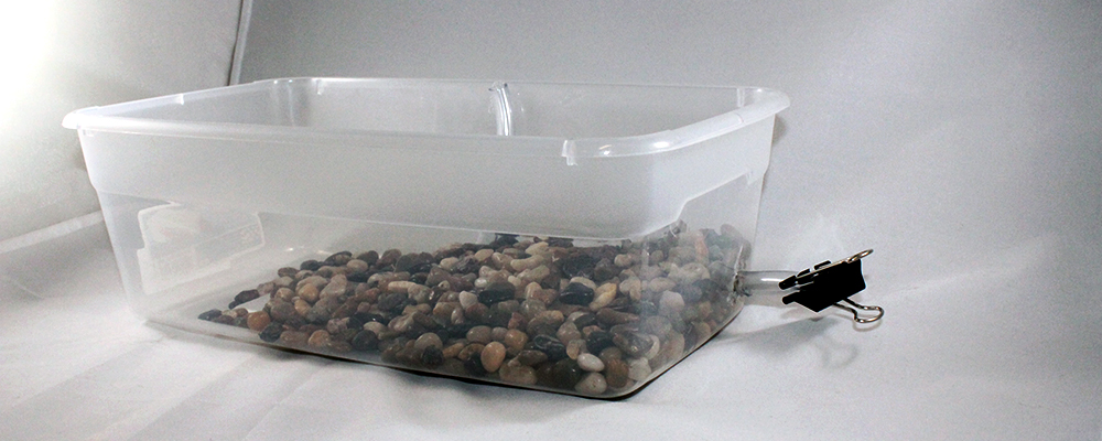 The container has a spout in the bottom corner, and it closed by a clip. Rocks are placed around the opening of the container. 